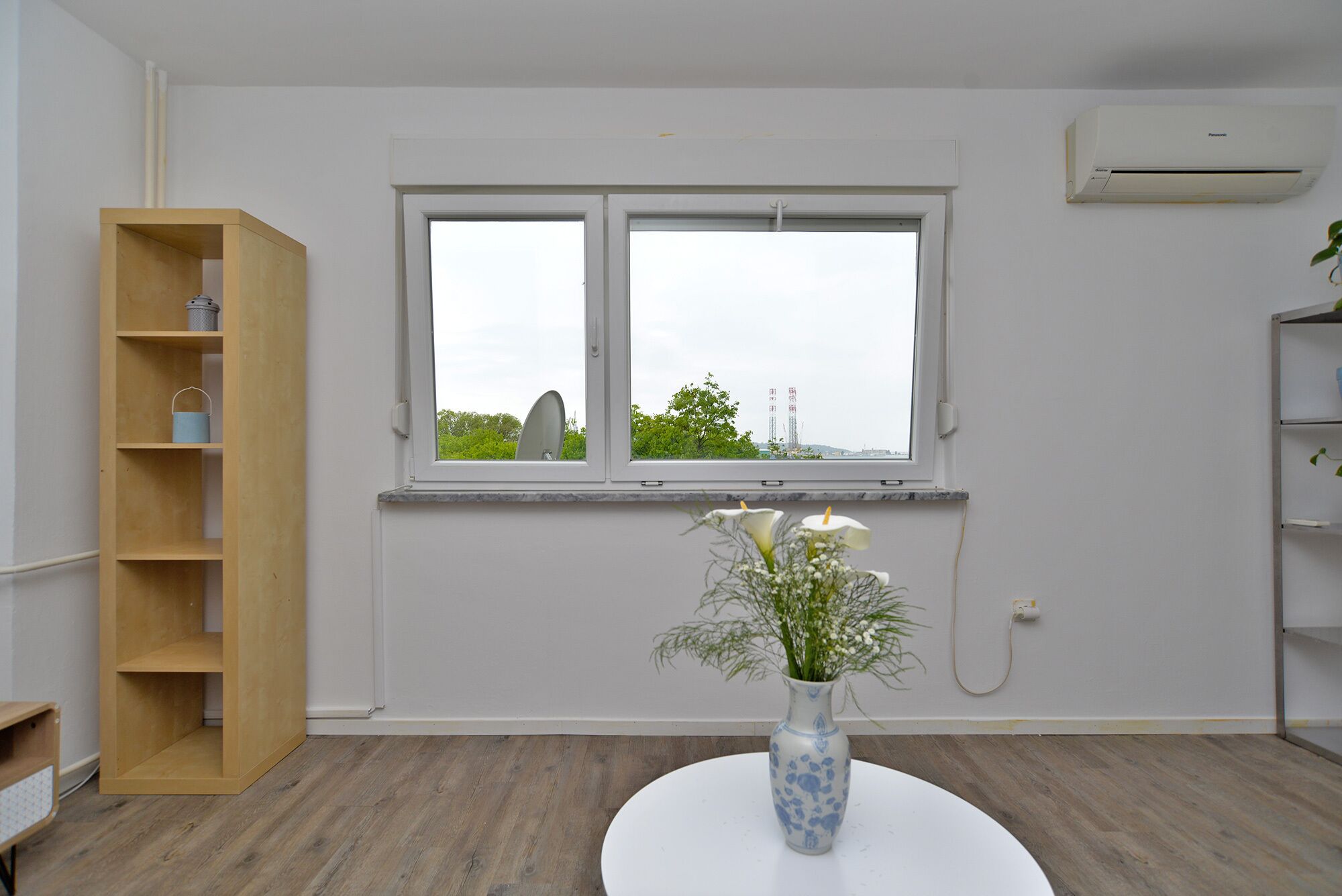 A room with a large window, a wooden bookshelf, a white round table with a vase, and an air conditioning unit.