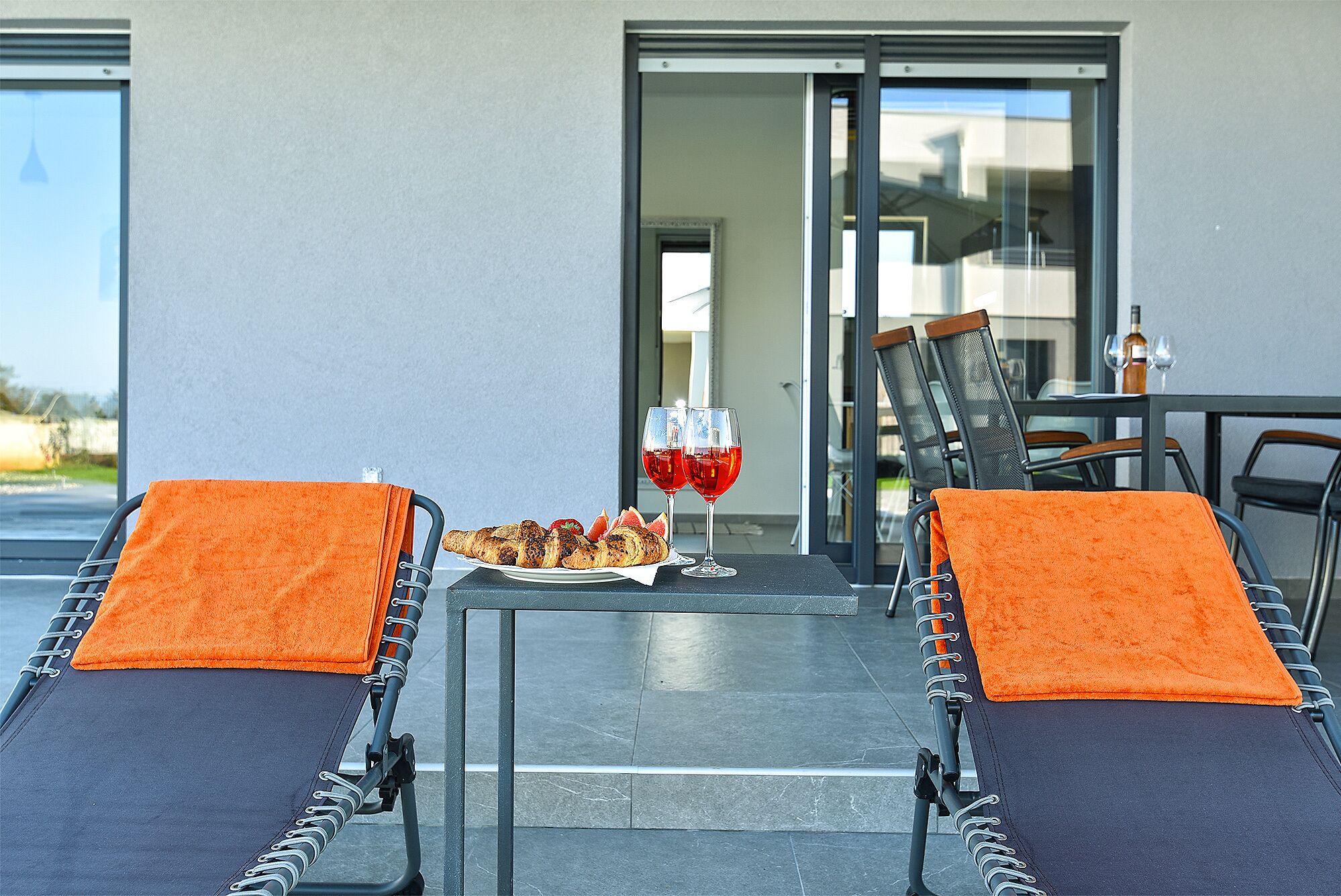 A modern outdoor seating area with two lounge chairs, a glass table, and a plate of food and drinks.