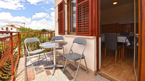 Un balcón con dos sillas y una mesa, un área de cocina visible a través de una puerta abierta, y vista de un edificio al fondo.