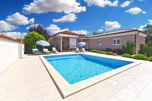 A swimming pool with lounge chairs and umbrellas, surrounded by a house with a tiled roof and a clear blue sky.