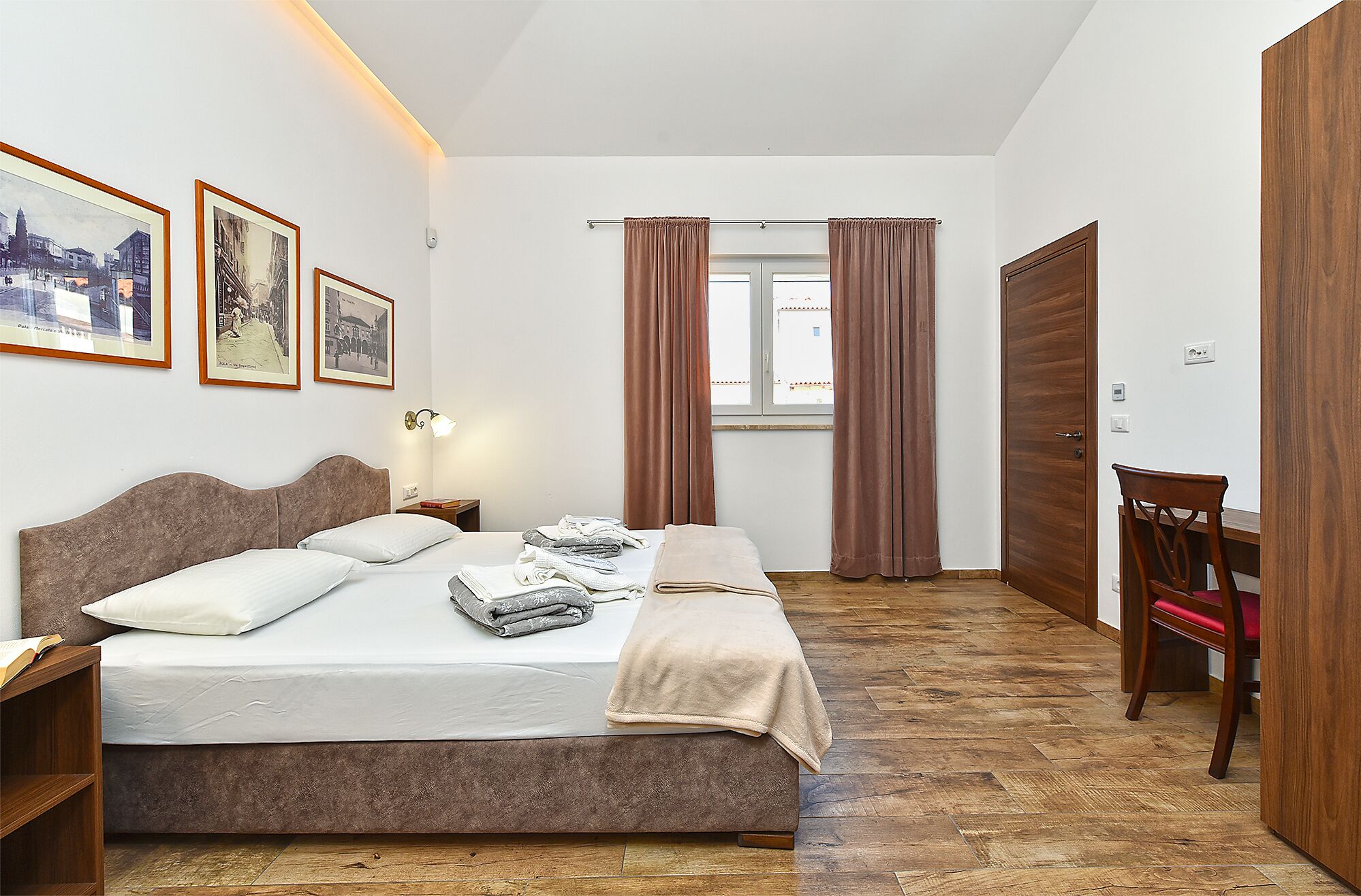 A bedroom with a bed, a bedside table, a lamp, a chair, a desk, a window with curtains, and framed pictures on the wall.