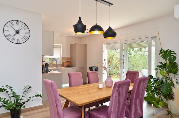 A modern kitchen with a wooden table and purple chairs, a clock on the wall, and a balcony with plants.