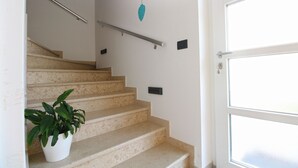 Un escalier avec des marches de couleur beige, un mur blanc, une plante en pot, une décoration suspendue et une porte avec des panneaux de verre dépoli.