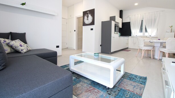 Un salon moderne avec un canapé gris, une table basse blanche et un coin cuisine doté de meubles blancs et d’un plan de travail noir.