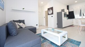 Un salon moderne avec un canapé gris, une table basse blanche et un coin cuisine doté de meubles blancs et d’un plan de travail noir.