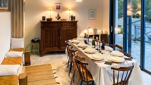 A dining area with a table set for four, wooden chairs, a wooden cabinet, and a view of the outdoors through large windows. - Maison Zenith - Five Bedroom Villa, Sleeps 10 (Beaurepaire)