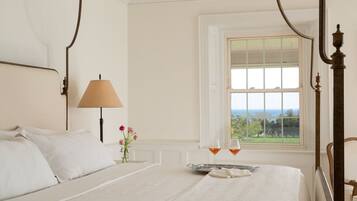 A bedroom with a four-poster bed, a bedside table with a lamp, a vase with flowers, and a window with a view.