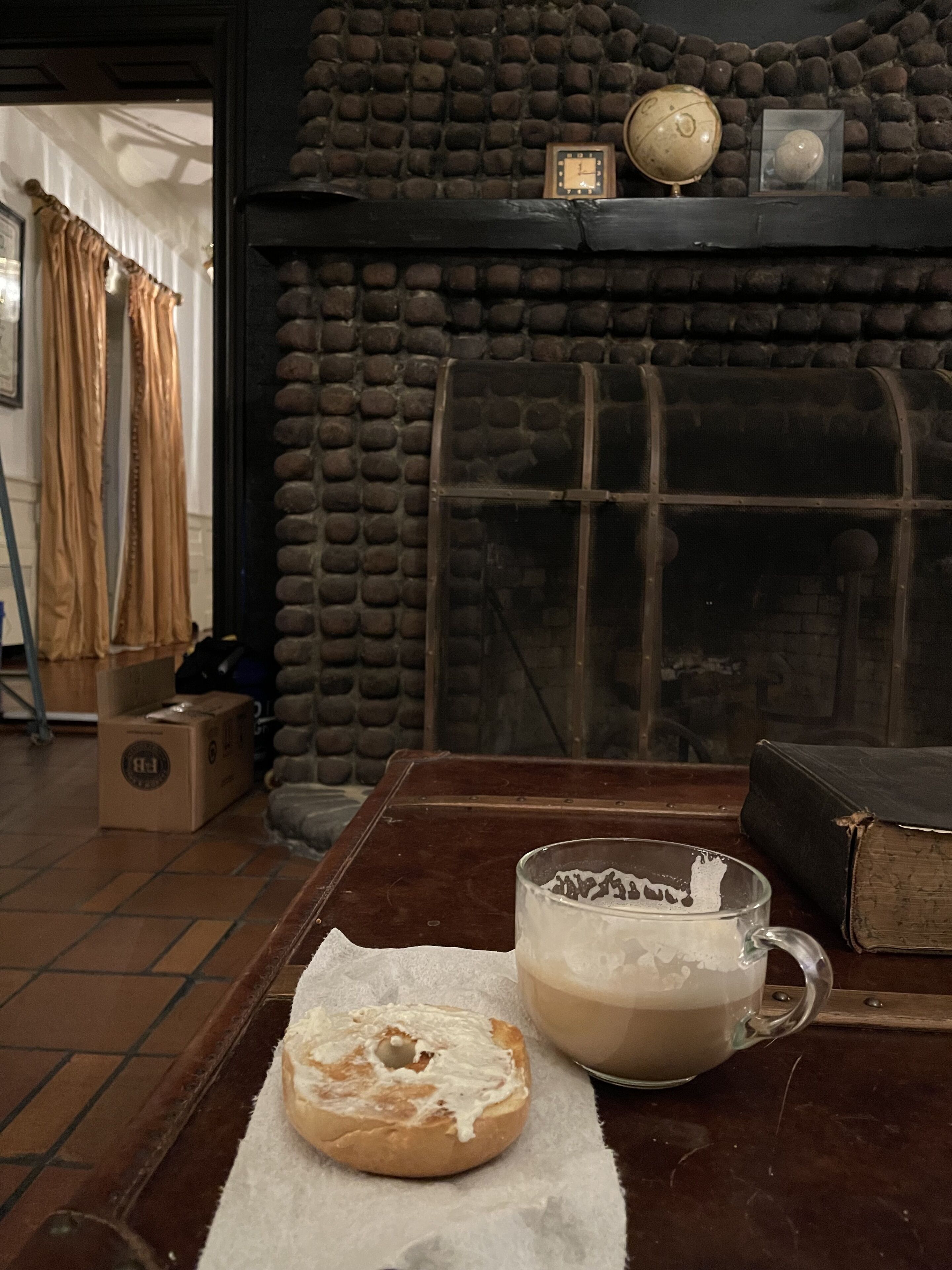 A rustic room with a stone wall, a wooden table with a mug and a bagel, and a bookshelf with books and a globe.