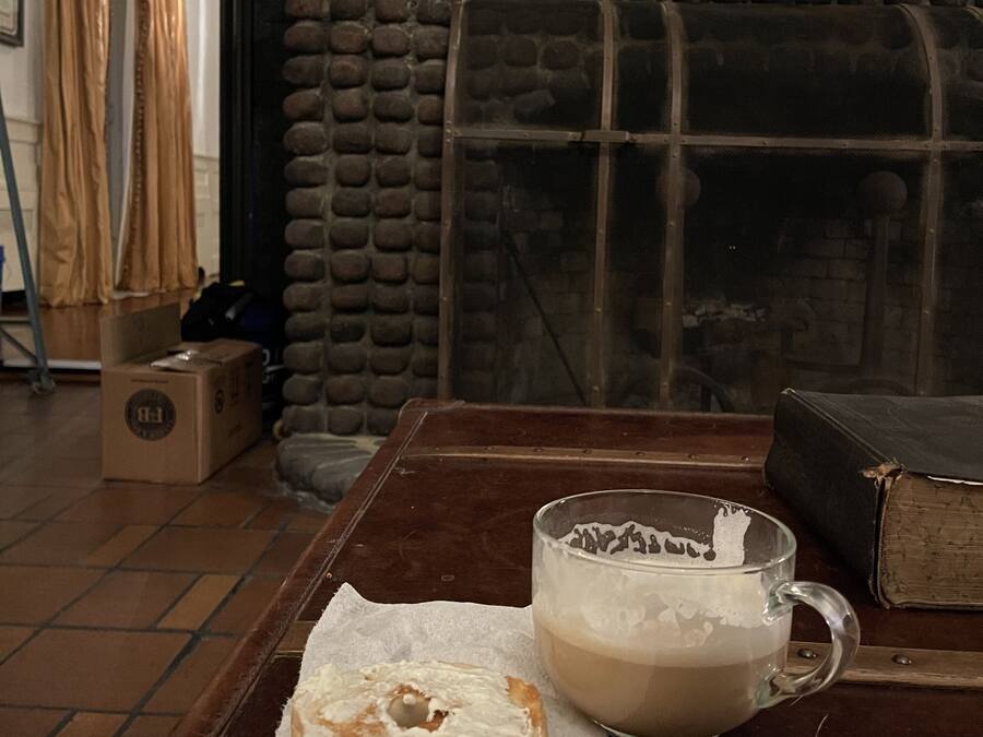 A rustic room with a stone wall, a wooden table with a mug and a bagel, and a bookshelf with books and a globe.