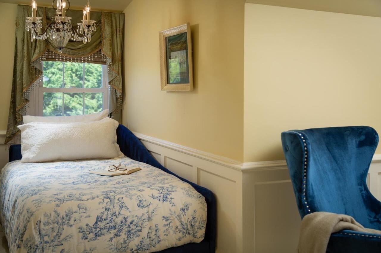 A bedroom with a bed, a blue chair, a chandelier, and a window with curtains.