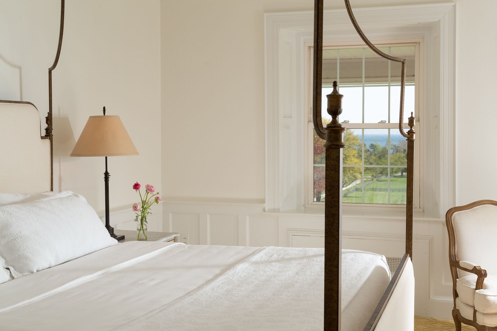 A bedroom with a four-poster bed, a bedside table with a lamp, a vase with flowers, and a view of a green landscape through a window.