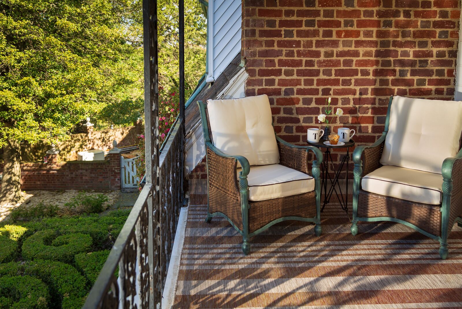 A balcony with wicker chairs and a small table, a brick wall, and greenery in the background.