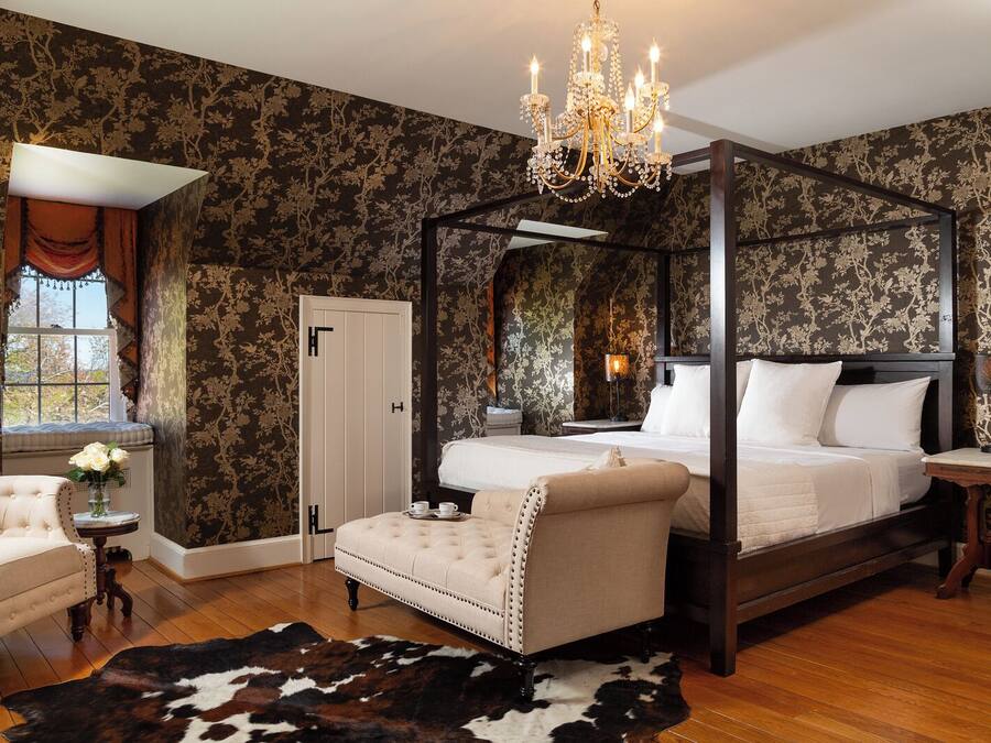 Uma cama com dossel e cortina, uma poltrona estofada, uma mesinha redonda com um vaso de flores e um tapete de couro sobre piso de madeira.