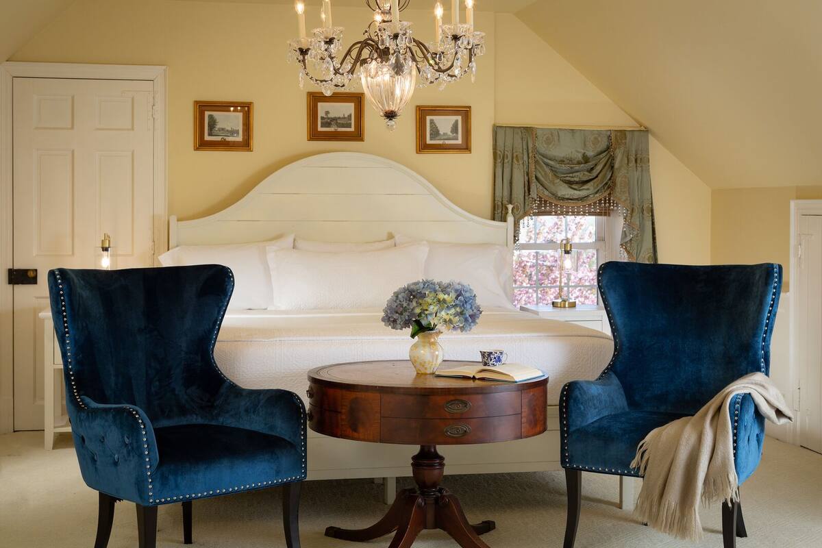Une chambre avec un grand lit, deux fauteuils en velours bleu, une table de chevet en bois, un lustre et des tableaux encadrés accrochés au mur.