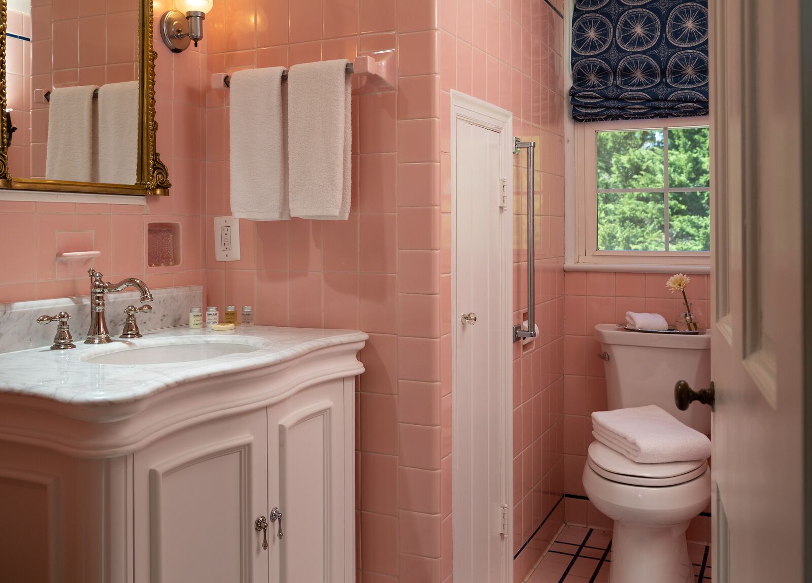 A bathroom with pink tiles, a white sink with gold faucets, a white toilet, and a window with a view of greenery.