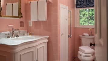 A bathroom with pink tiles, a white sink with gold faucets, a white toilet, and a window with a view of greenery.