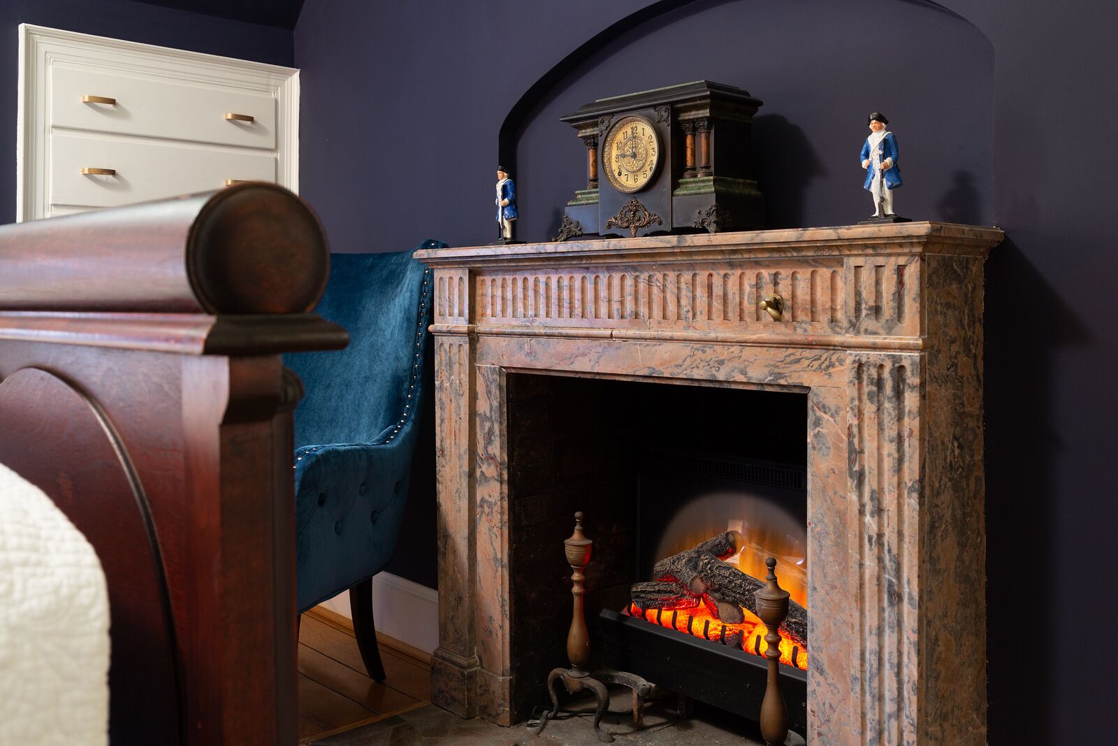 A fireplace with a clock and figurines on top, a blue armchair, and a wooden chair with a cushion.