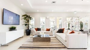 A modern living room with a white sofa set, a coffee table, and a large TV on the wall. There are potted plants and a view of the outside through large windows. - 4 Suites Villa 5 min to Disney (Kissimmee)