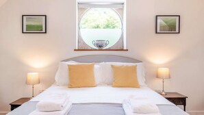 Cama bem arrumada com almofadas brancas e amarelas, duas luminárias de cabeceira e uma janela com vista para o verde.