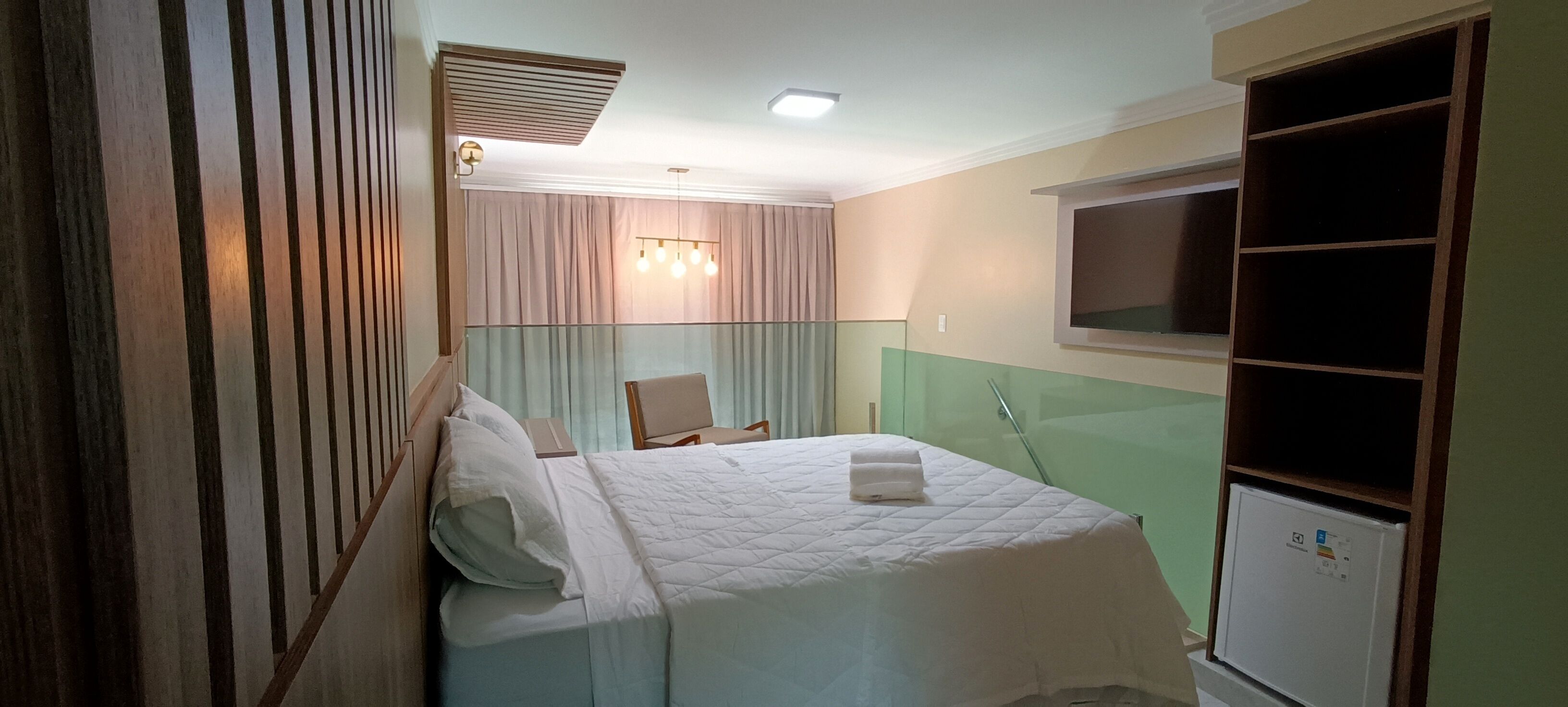 Una camera d'albergo con un letto, una sedia, uno specchio e un piccolo frigorifero.