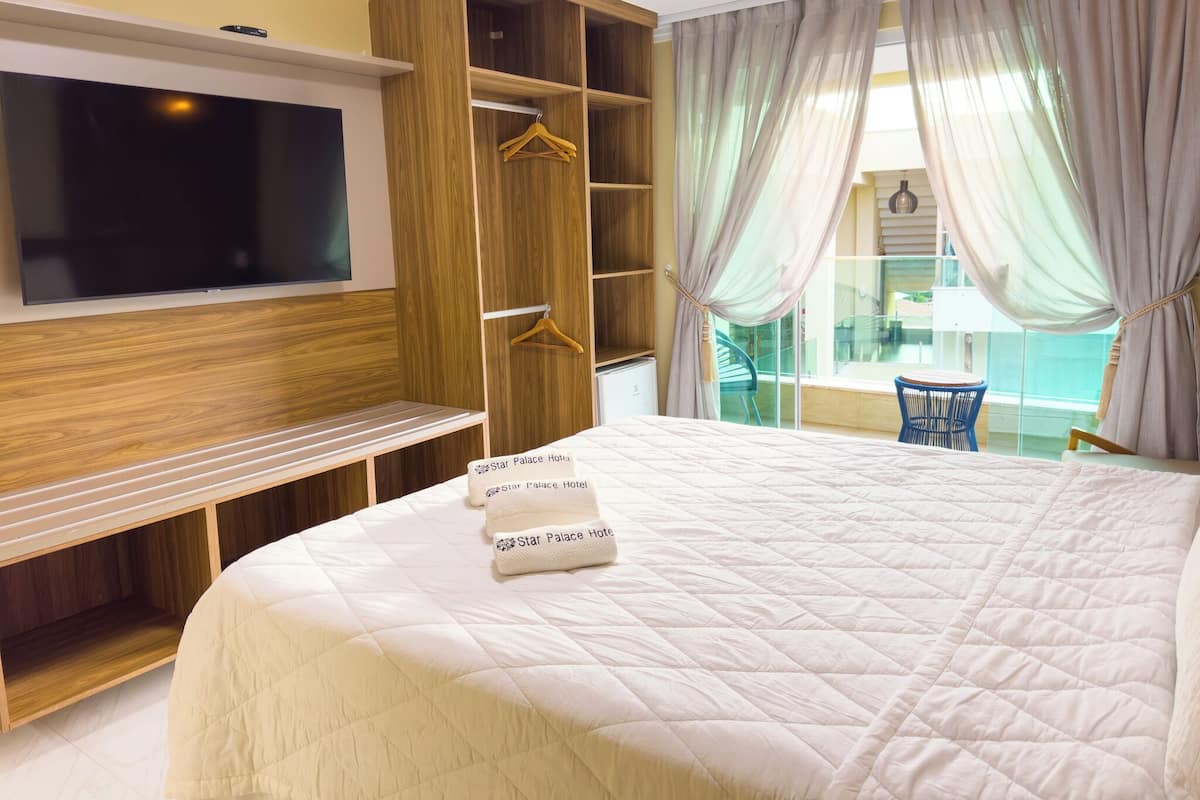 A hotel room with a large bed, a TV mounted on the wall, wooden furniture, and a balcony with a view.