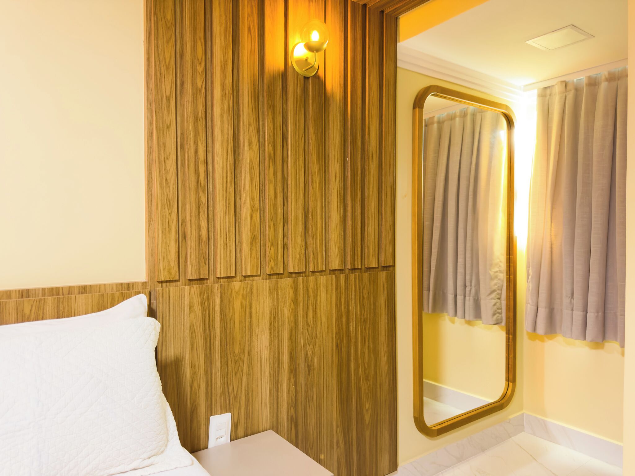 A hotel room with a wooden panel wall, a bed with white bedding, a bedside table, a wall-mounted mirror, and a window with curtains.