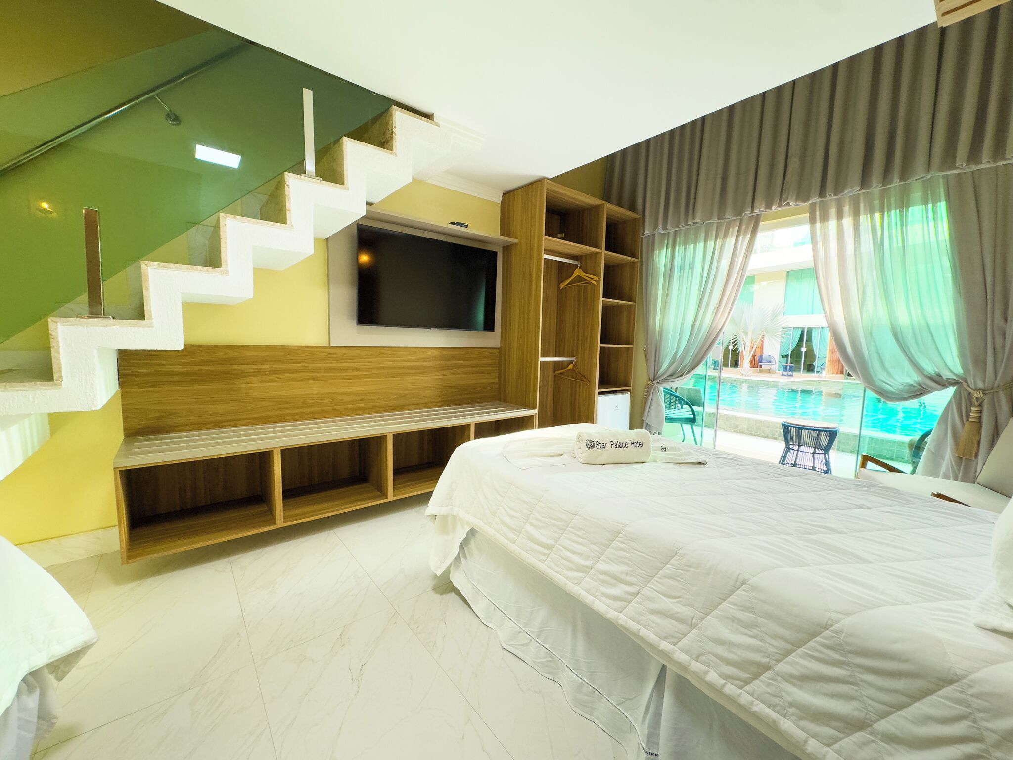 A modern hotel room with a large bed, a flat-screen TV mounted on the wall, a wooden bench, and a view of a swimming pool.