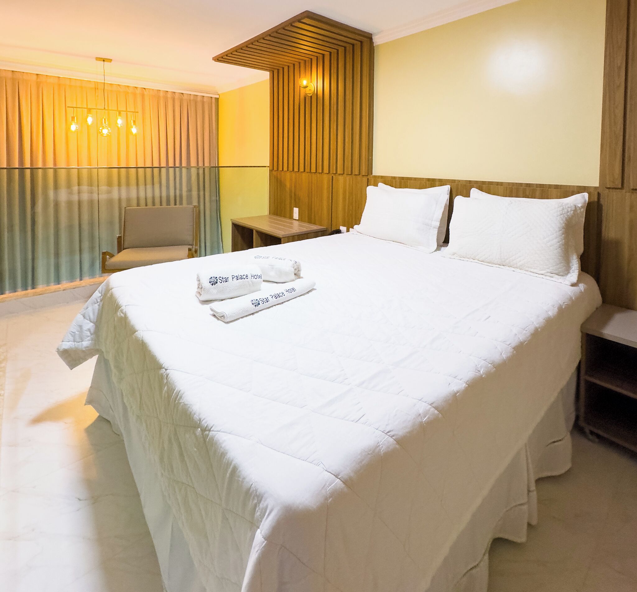 A hotel room with a large bed, a chair, a small table, and a wall-mounted light fixture.
