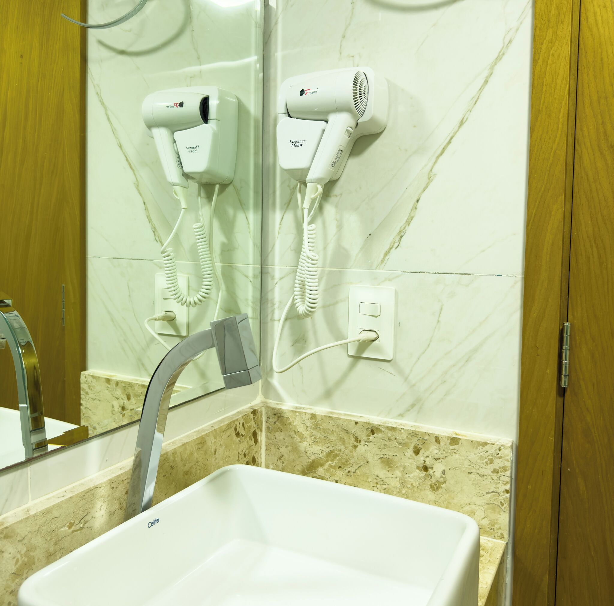 A bathroom with a marble wall, a sink, and two hair dryers mounted on the wall.