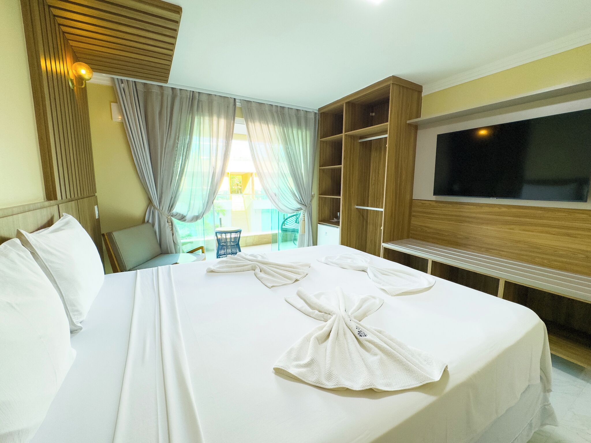 A hotel room with a large bed, a TV, a wooden wardrobe, and a balcony with a chair.