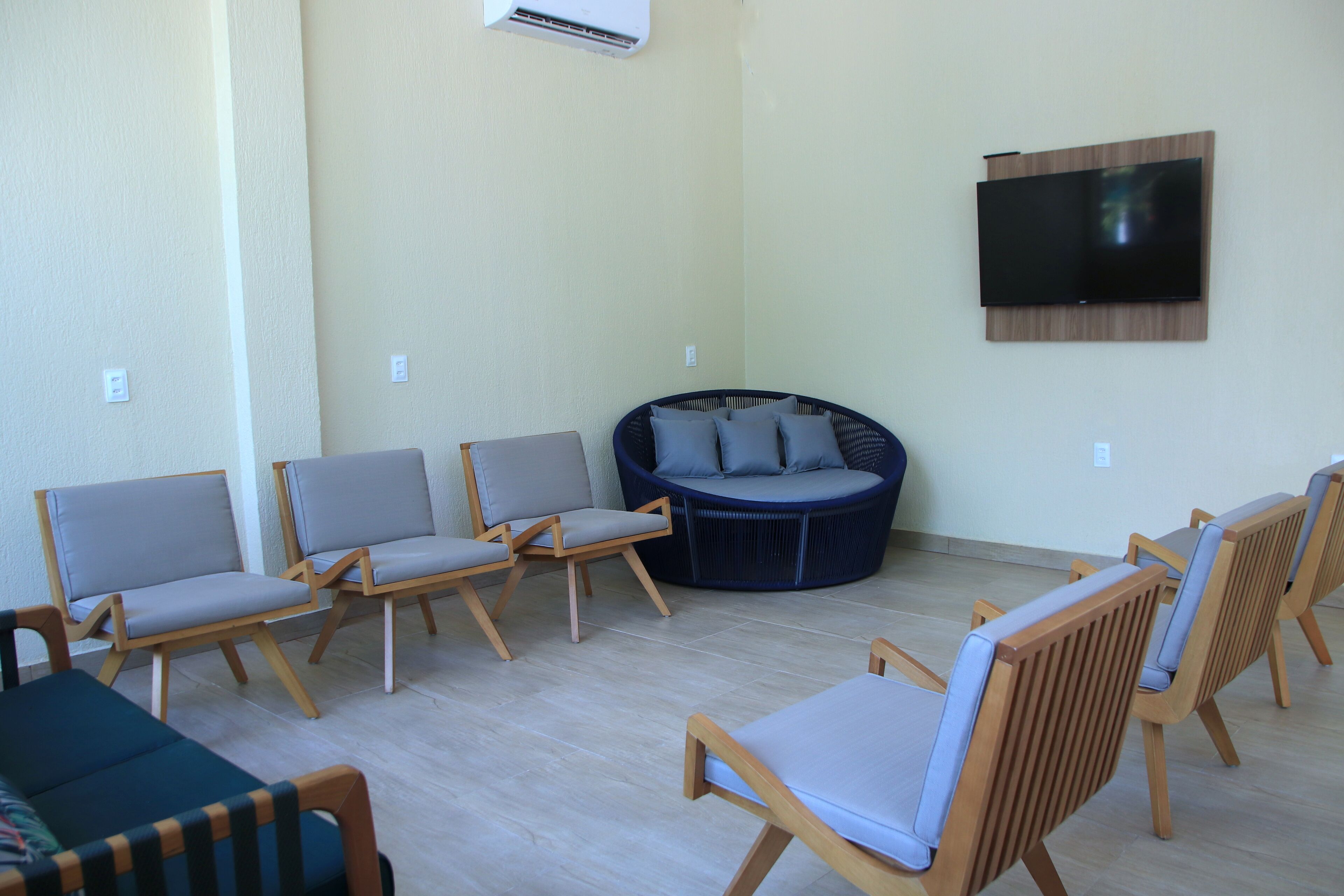 A room with a flat-screen TV mounted on the wall, a round seating area with cushions, and several wooden chairs with cushions.