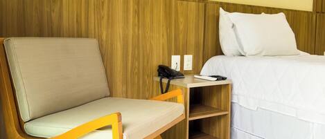 A hotel room with a wooden headboard, a bed with white bedding, a bedside table with a phone and a lamp, and a chair with a light cushion.