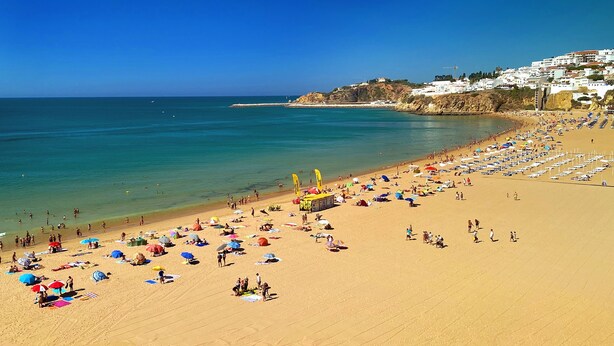 A beach with numerous umbrellas and sunbeds, a clear blue sky, and a distant view of a town on a hill. - B&B Albufeira City Room Junior with Private Bathroom (Albufeira)