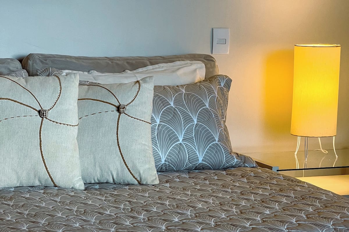 A neatly made bed with patterned bedding and decorative pillows, a bedside lamp, and a wall-mounted light switch.