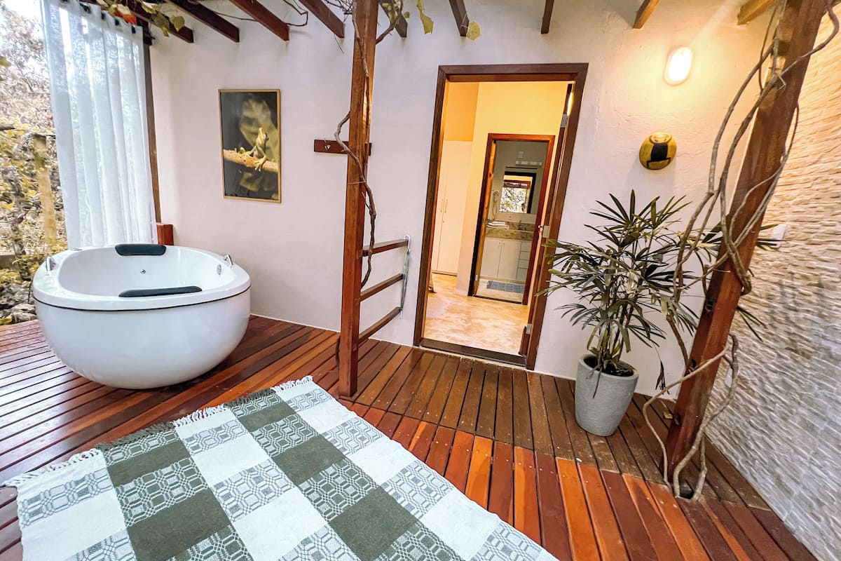 A bathroom with a large bathtub, wooden flooring, a checkered rug, and a potted plant.