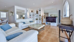 A modern living room with a white sofa, a glass coffee table, and a flat-screen TV. There are shelves with decorative items and a large mirror. - Apartment Nautica Bay Moraira (Moraira)