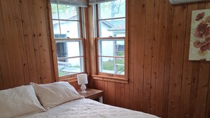 A bedroom with a large bed, wooden walls, a window with blinds, a nightstand with a lamp, and an air conditioning unit.