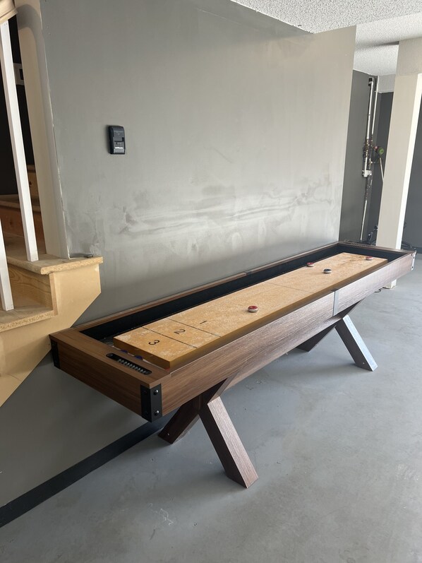 A shuffleboard table with a wooden frame and metal legs, positioned against a plain wall with a visible electrical outlet.