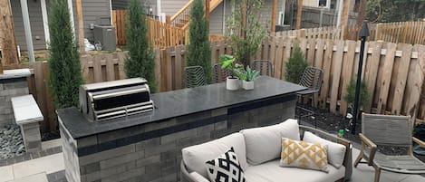 An outdoor patio area with a built-in grill, a cushioned seating area, and a wooden fence.