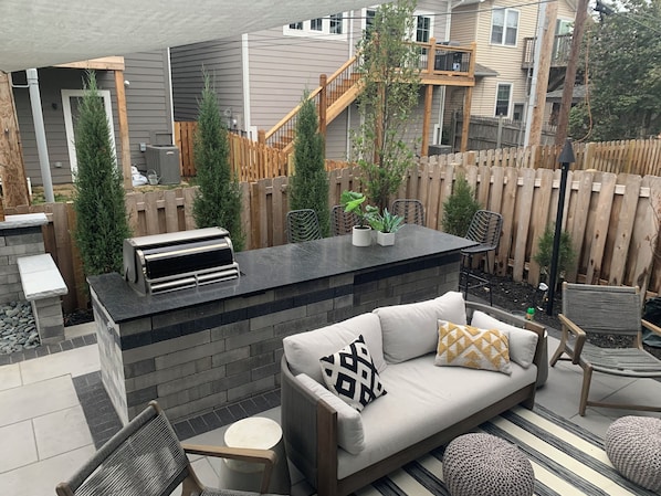 Un patio exterior con una parrilla integrada, una zona de estar con cojines y una valla de madera.