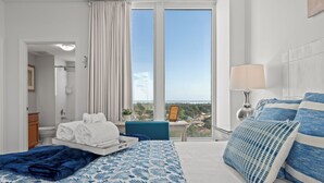 Ein Hotelzimmer mit Bett, Fenster mit Vorhängen, Nachttisch mit Lampe und Blick auf die Stadt.