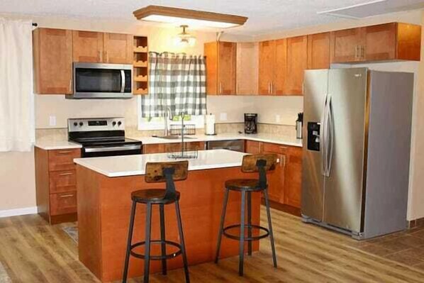 A kitchen with wooden cabinets, a stainless steel refrigerator, a microwave, a stove, and two bar stools.