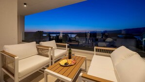 Un balcon doté de sièges blancs et confortables, une table en bois avec une bouteille et des verres, et une vue imprenable sur un coucher de soleil se reflétant sur une étendue d’eau.