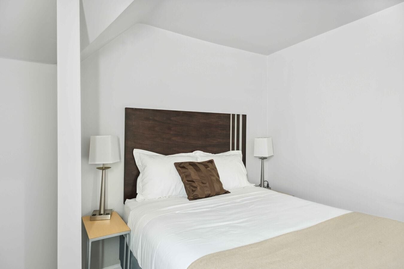 A hotel room with a bed, two bedside lamps, a wooden headboard, and a beige throw blanket.