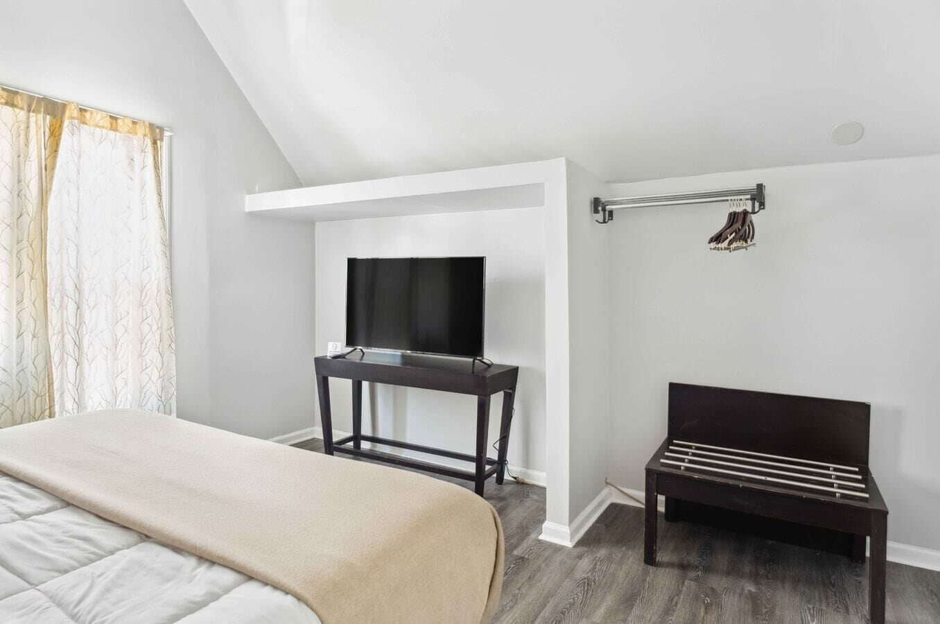 A modern hotel room with a bed, a TV on a stand, a coat rack, and a wooden bench.