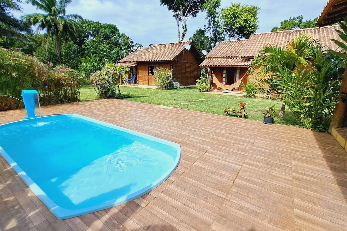 Uma piscina com deck de madeira, cercada por vegetação exuberante e duas casas rústicas com telhados de azulejo.