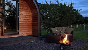 A cozy outdoor seating area with a fire pit, a sofa, and a table set for two, adjacent to a wooden building with a glass door.