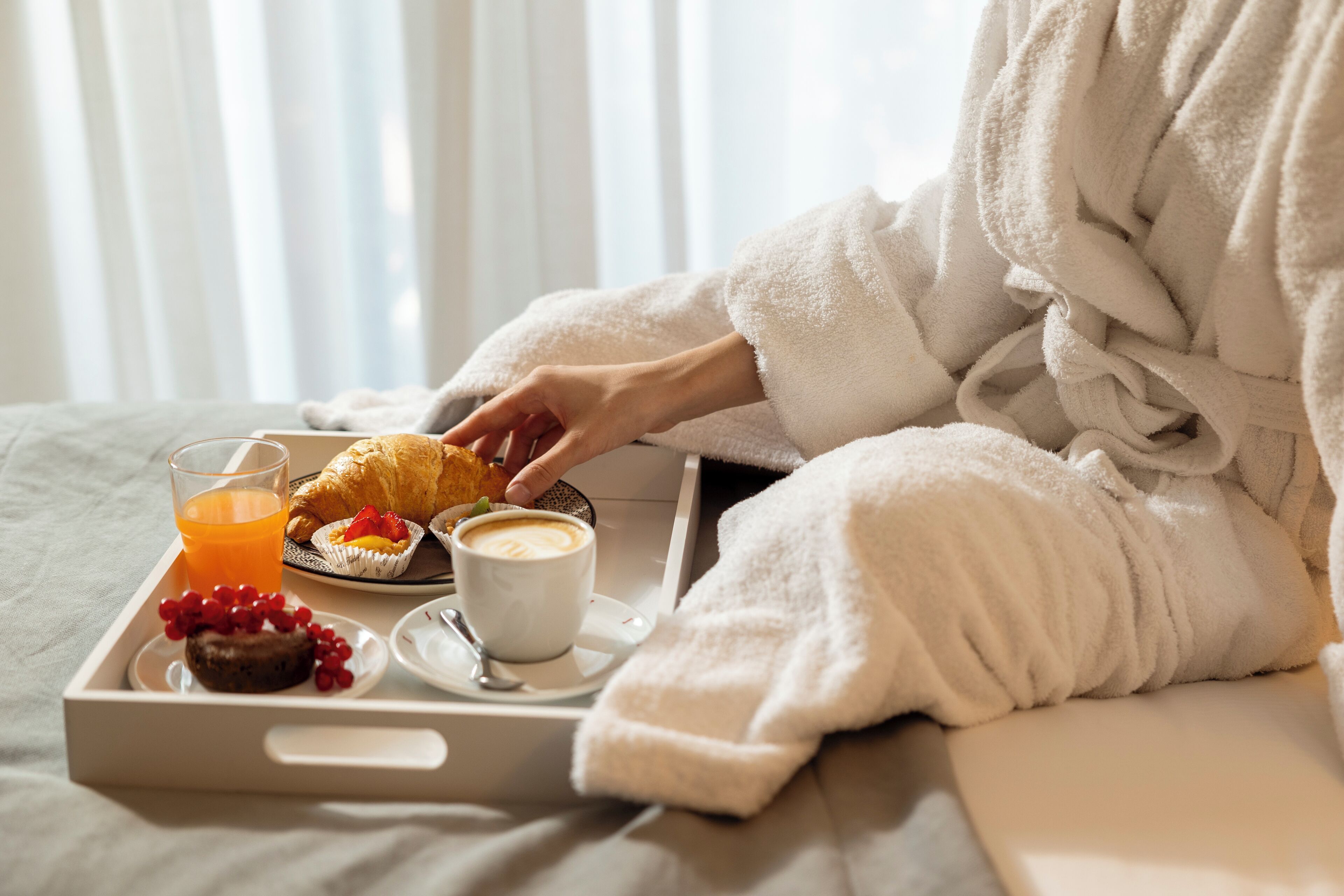 Une personne vêtue d’un peignoir blanc est assise sur un lit, et devant elle se trouve un plateau contenant un croissant, un muffin, une tasse de café et un verre de jus d’orange.