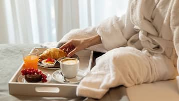 Une personne vêtue d’un peignoir blanc est assise sur un lit, et devant elle se trouve un plateau contenant un croissant, un muffin, une tasse de café et un verre de jus d’orange.