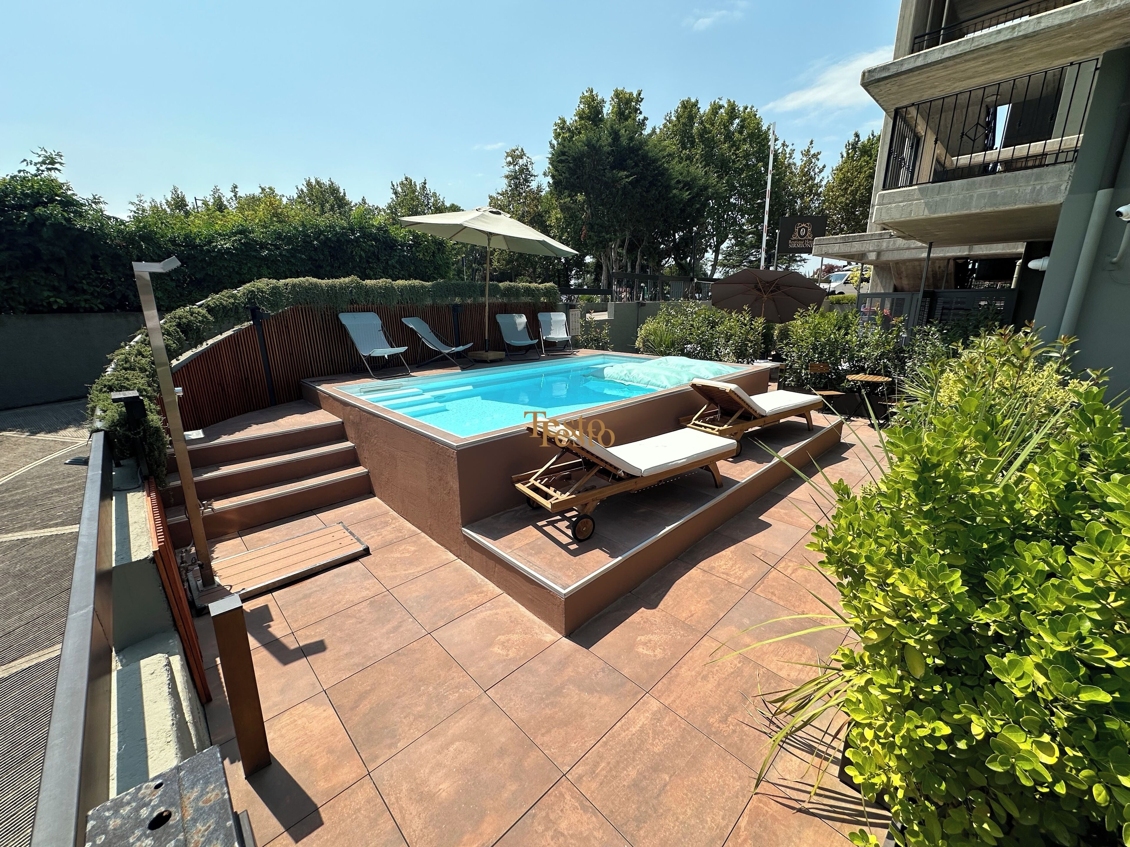 A rectangular swimming pool with a tiled deck, surrounded by lounge chairs and a pergola with an umbrella.